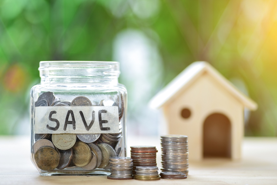 A jar with coins in it with tape on the front that says "SAVE". Next to the jar are several stacks of coins. Behind the jar is a out of focus mini-house